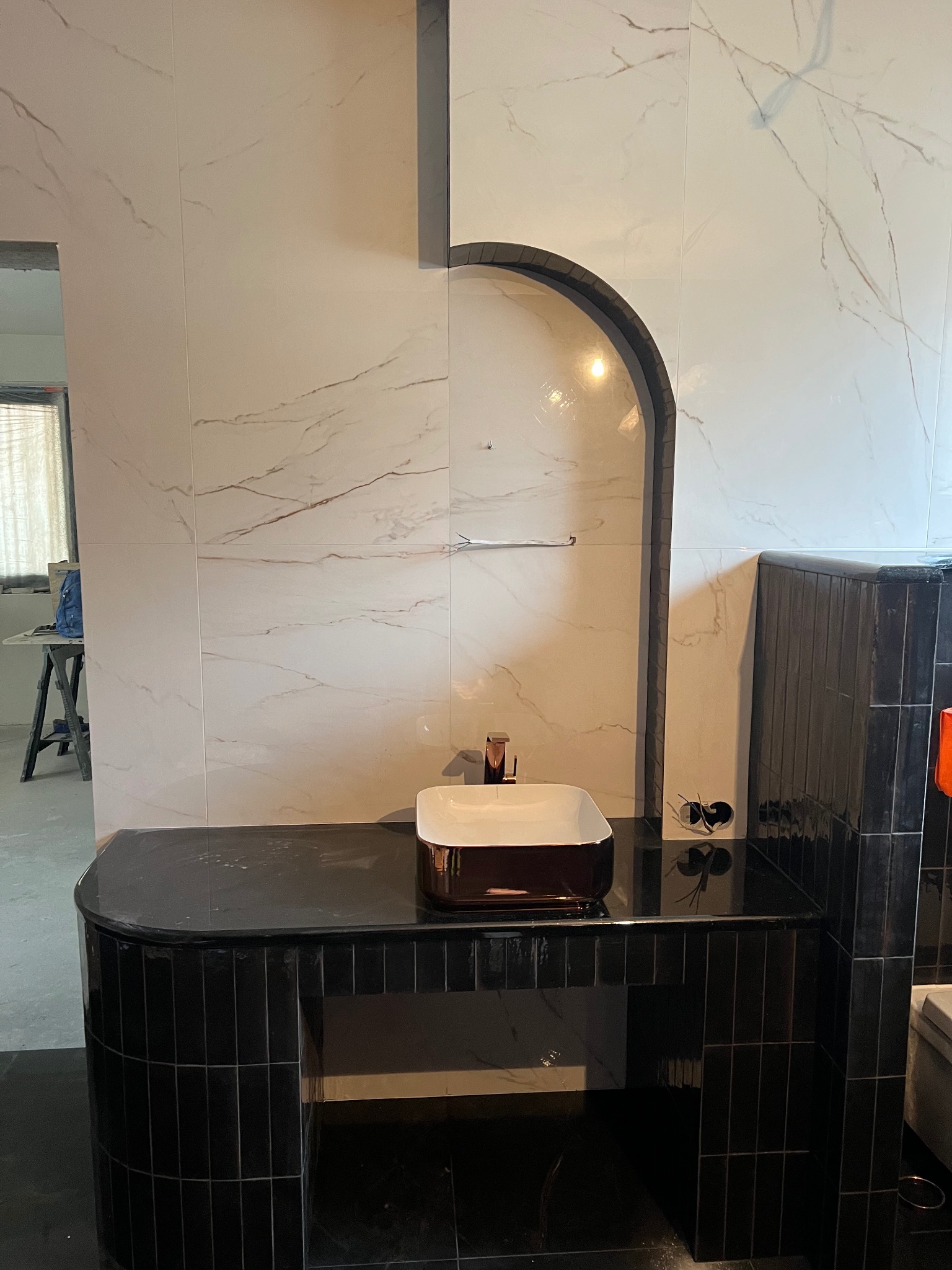 Wykończona łazienka z umywalką nablatową w kolorze brązowo-białym, czarnym blatem i ścianami wyłożonymi płytkami imitującymi marmur z brązowymi żyłkami. Widoczne gniazdka elektryczne bez osprzętu.