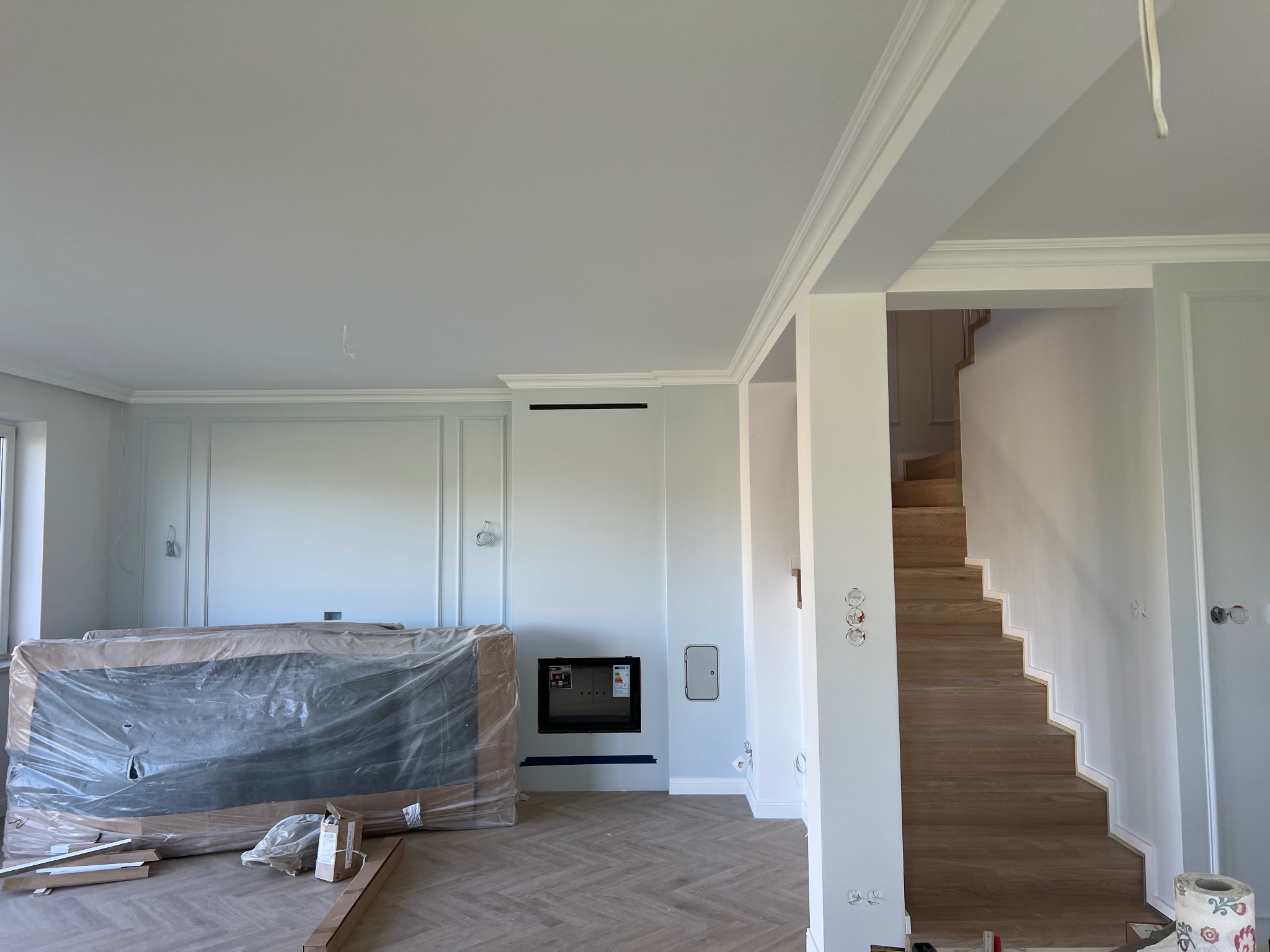 Wnętrze w trakcie wykończenia z parkietem w jodełkę, białą sztukaterią, schodami drewnianymi i kominkiem, zabezpieczony mebel w folii.