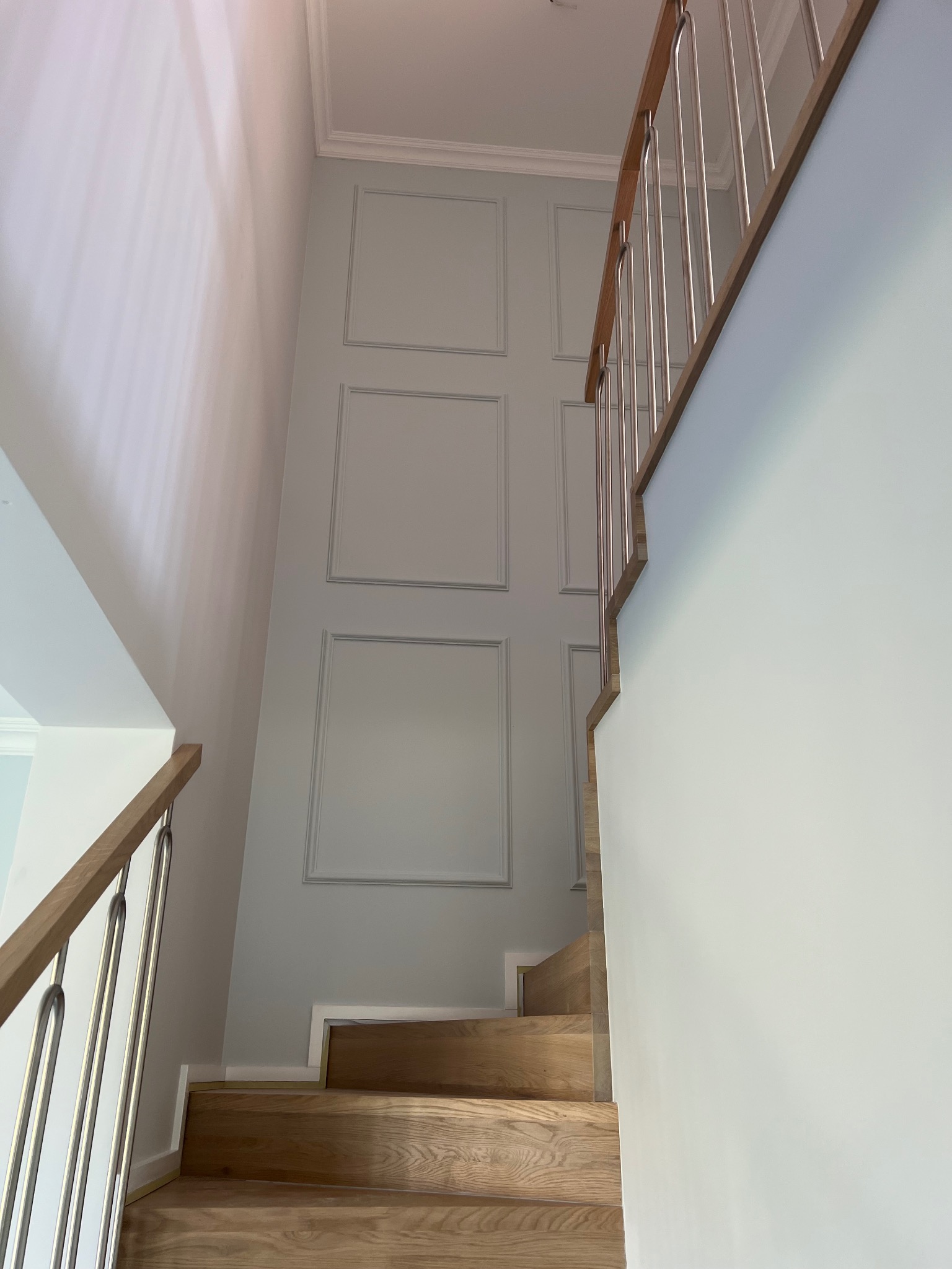 Wnętrze klatki schodowej z drewnianymi schodami, metalową balustradą i ścianą w pastelowym odcieniu z dekoracyjnymi panelami.