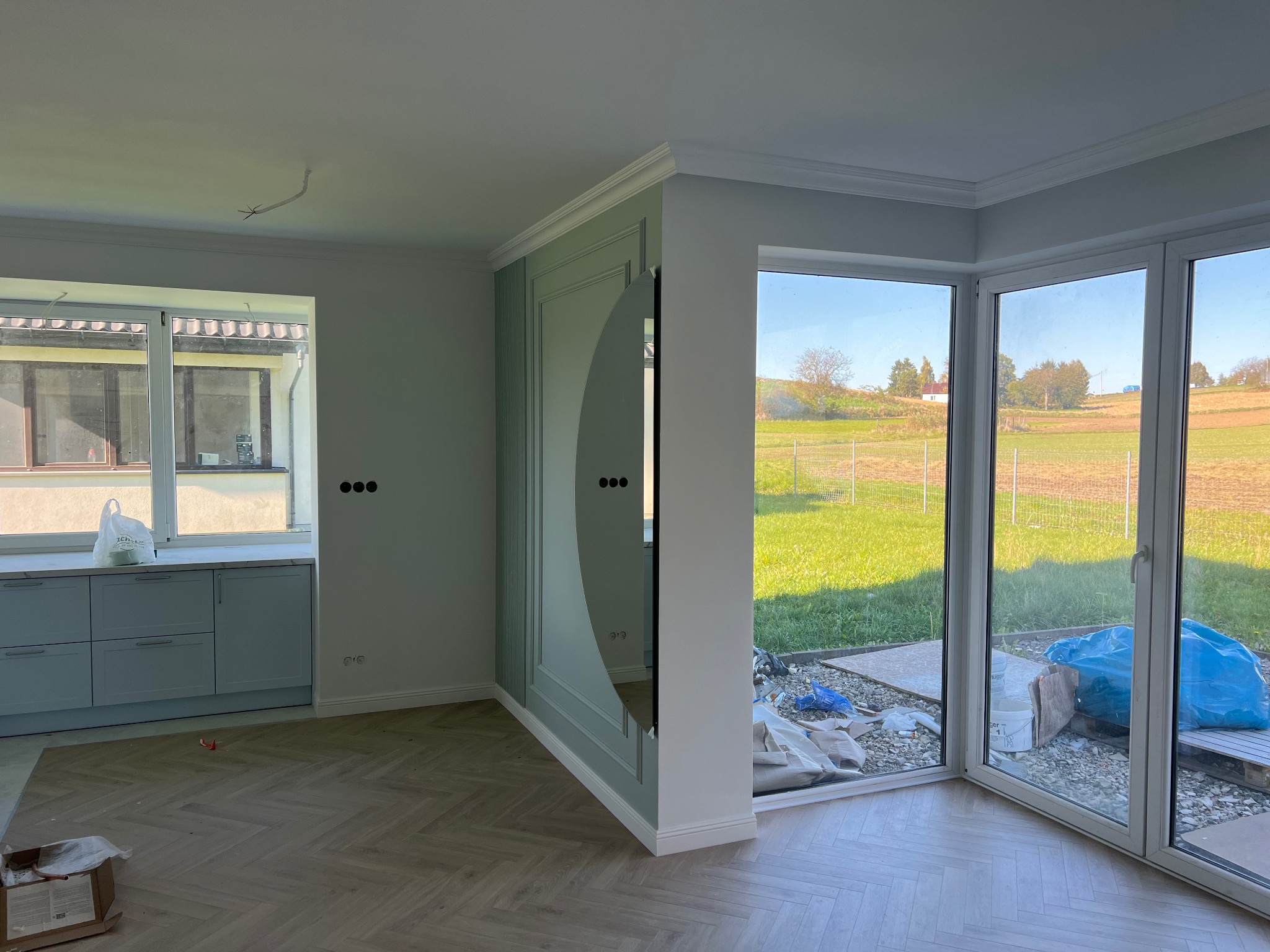 Wnętrze w trakcie wykańczania: parkiet ułożony w jodełkę, jasne ściany z dekoracyjną sztukaterią w odcieniach zieleni, widoczne okno z widokiem na zewnątrz i fragment zabudowy kuchennej.