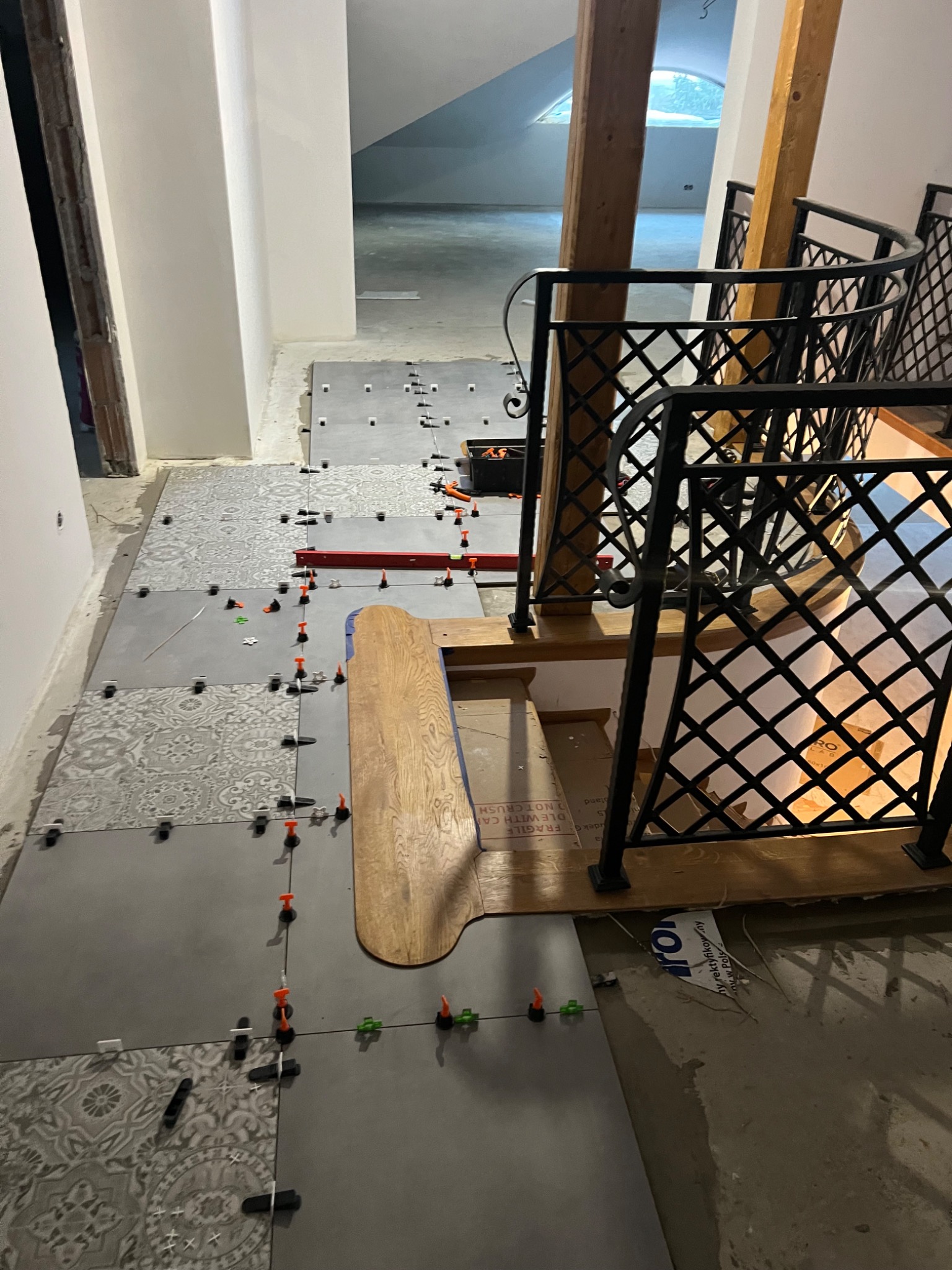 Układanie płytek podłogowych na schodach i korytarzu, widoczne poziomice, kliny dystansowe i wzorzyste płytki w szarościach, balustrada schodowa z metaloplastyki