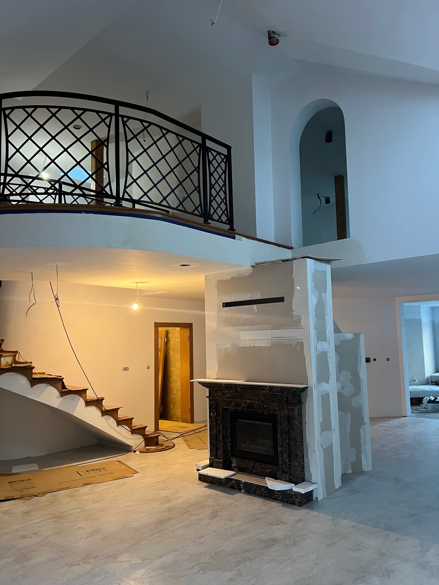 Dwupoziomowe wnętrze w trakcie remontu: marmurowy kominek, schody z drewnianymi stopniami, balkon z ozdobną balustradą, ściany przygotowane do malowania, widoczne kable i pojedyncza żarówka.