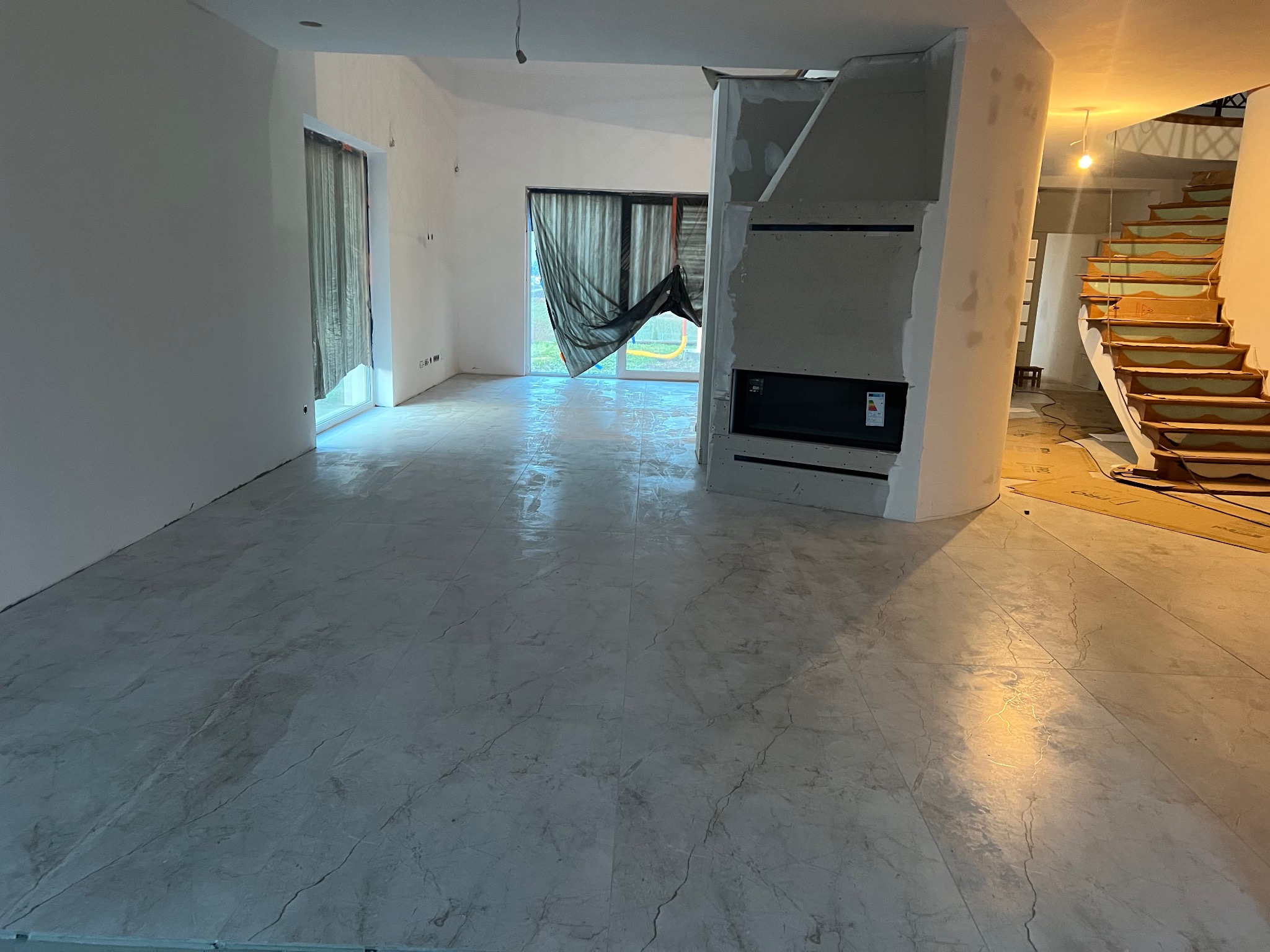 Wnętrze domu w trakcie prac wykończeniowych: marmurowe płytki podłogowe, surowe ściany przygotowane pod malowanie, obudowa kominka z płyt kartonowo-gipsowych, zabezpieczone schody z drewnianymi...