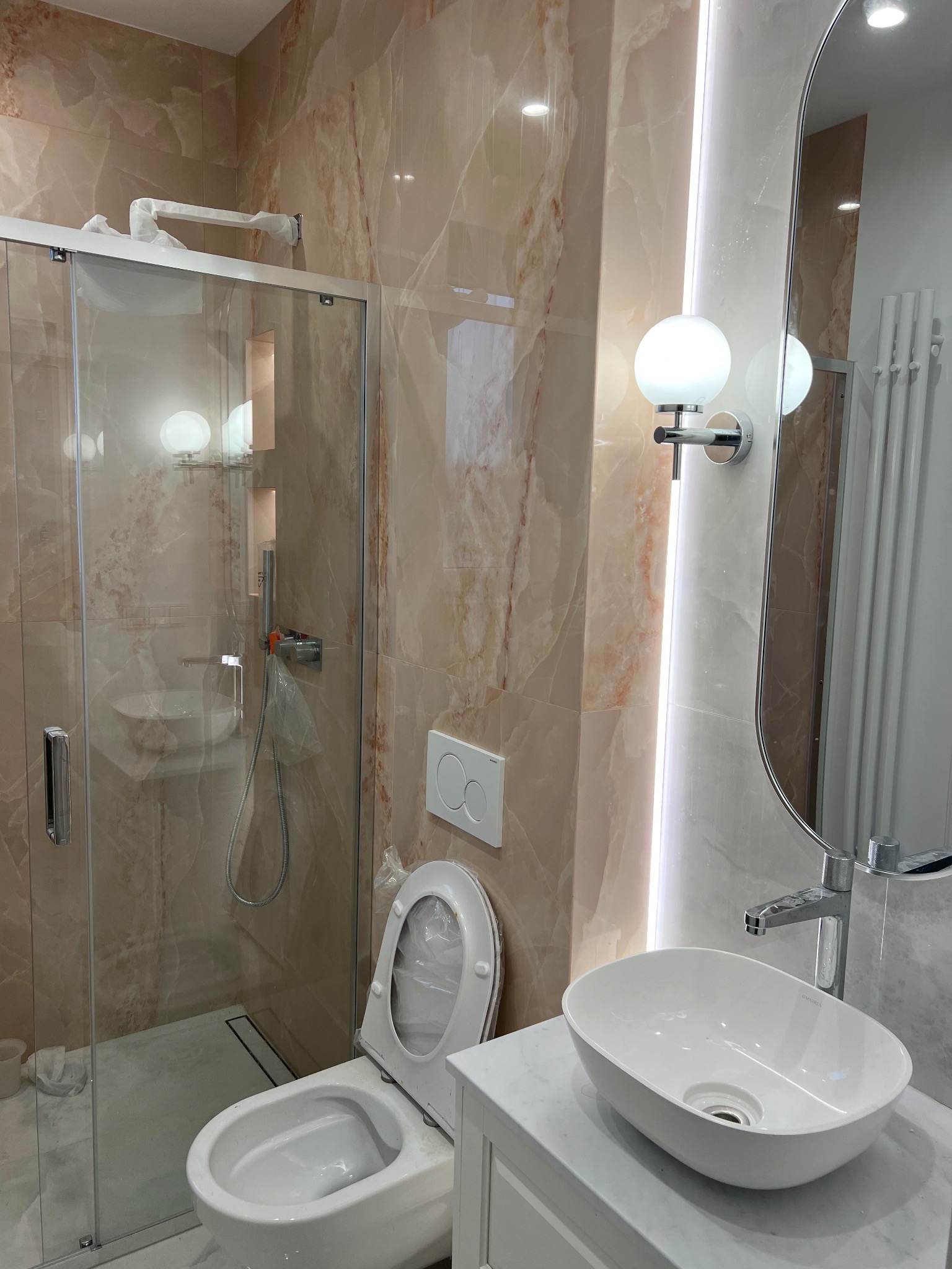 Wnętrze łazienki w trakcie wykańczania, z kabiną prysznicową z elementami zabezpieczonymi folią, umywalką nablatową i toaletą z otwartą klapą, na tle marmurowych płytek.