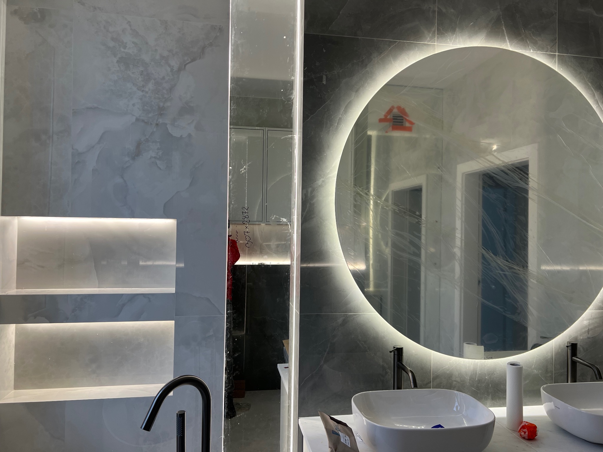 Nowoczesna łazienka z marmurowymi płytkami, okrągłym lustrem z podświetleniem LED, dwiema białymi umywalkami nablatowymi i czarną armaturą, częściowo zabezpieczona folią ochronną po remoncie.