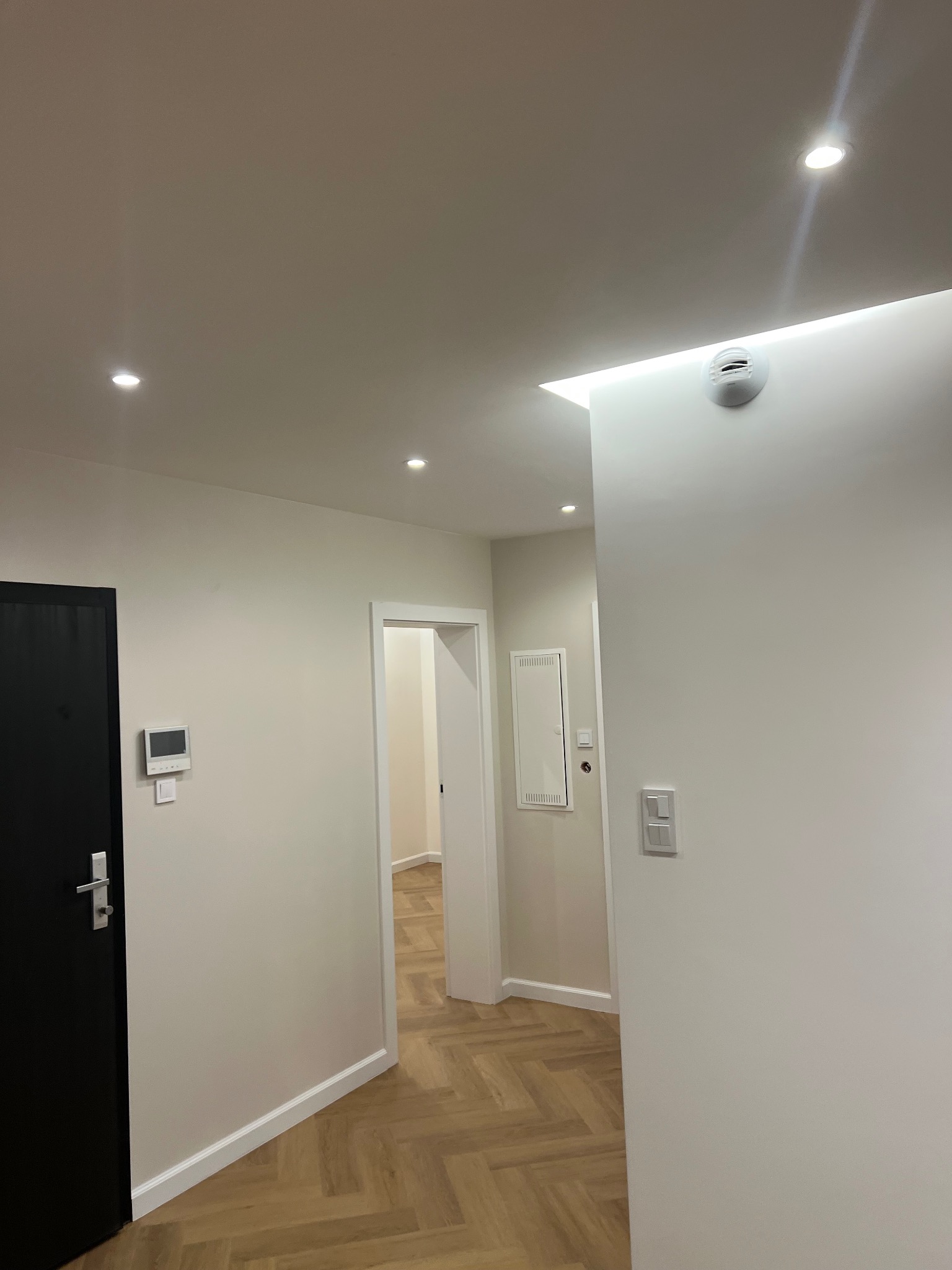 Korytarz po remoncie z parkietem w jodełkę, świeżo pomalowanymi ścianami w odcieniach bieli, czarnymi drzwiami i nowoczesnym oświetleniem punktowym oraz listwami przypodłogowymi.