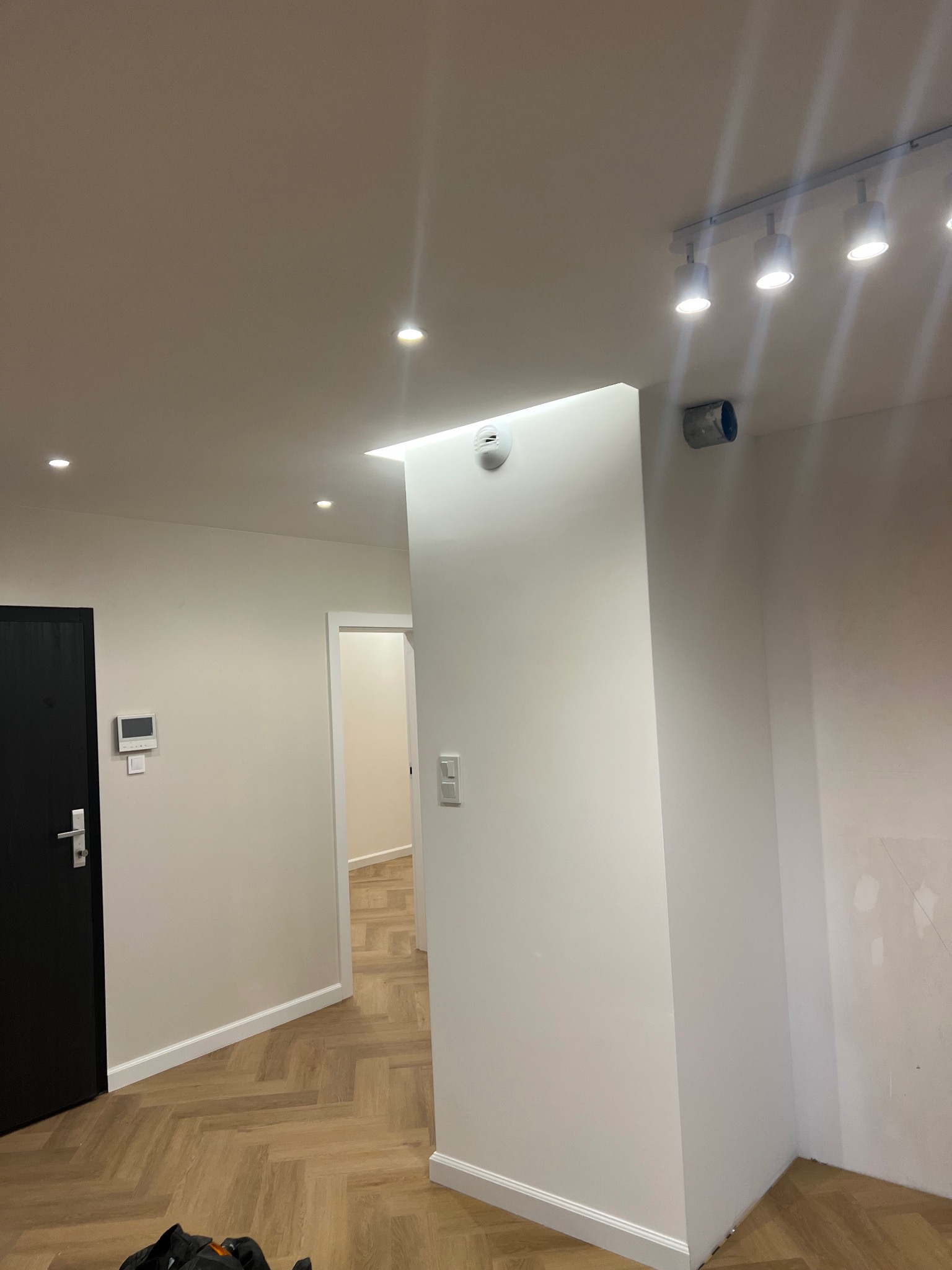 Wnętrze po remoncie: świeżo pomalowane, białe ściany z listwami przypodłogowymi, parkiet ułożony w jodełkę, czarne drzwi wejściowe i oświetlenie punktowe na suficie.