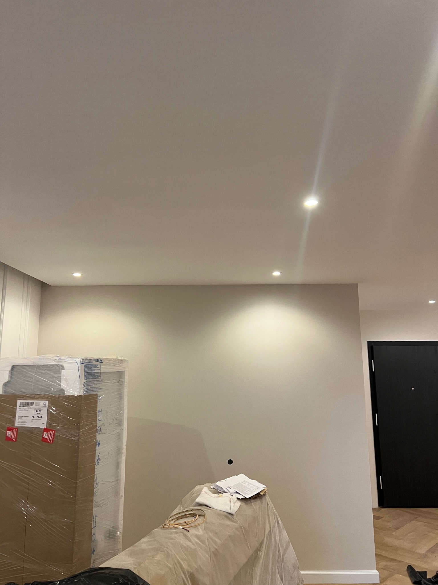 Wnętrze w trakcie prac wykończeniowych: gładka, beżowa ściana z trzema punktowymi lampami sufitowymi, zabezpieczone meble w folii stretch i kartonach, widoczny fragment drewnianej podłogi i czarne...