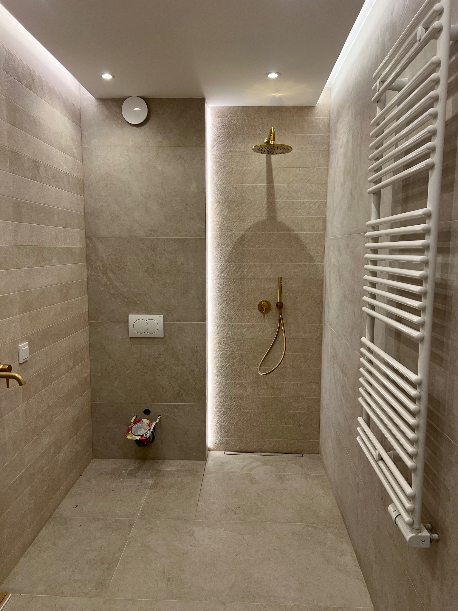 Nowoczesna łazienka z beżowymi płytkami, złotą armaturą prysznicową i białym grzejnikiem drabinkowym, widoczne przygotowania do montażu umywalki.