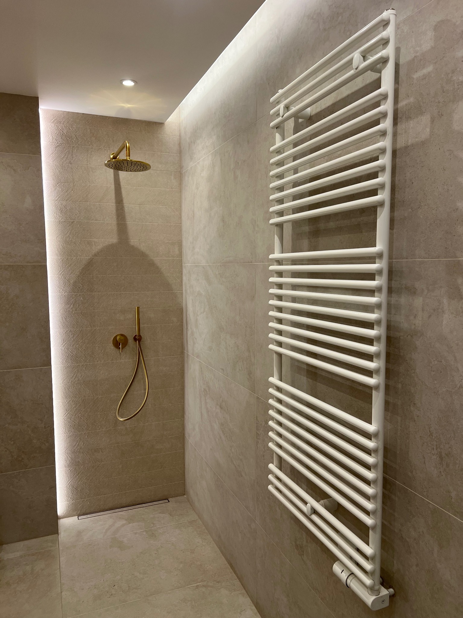 Nowoczesna kabina prysznicowa z złotą armaturą i białym grzejnikiem drabinkowym na beżowych płytkach.