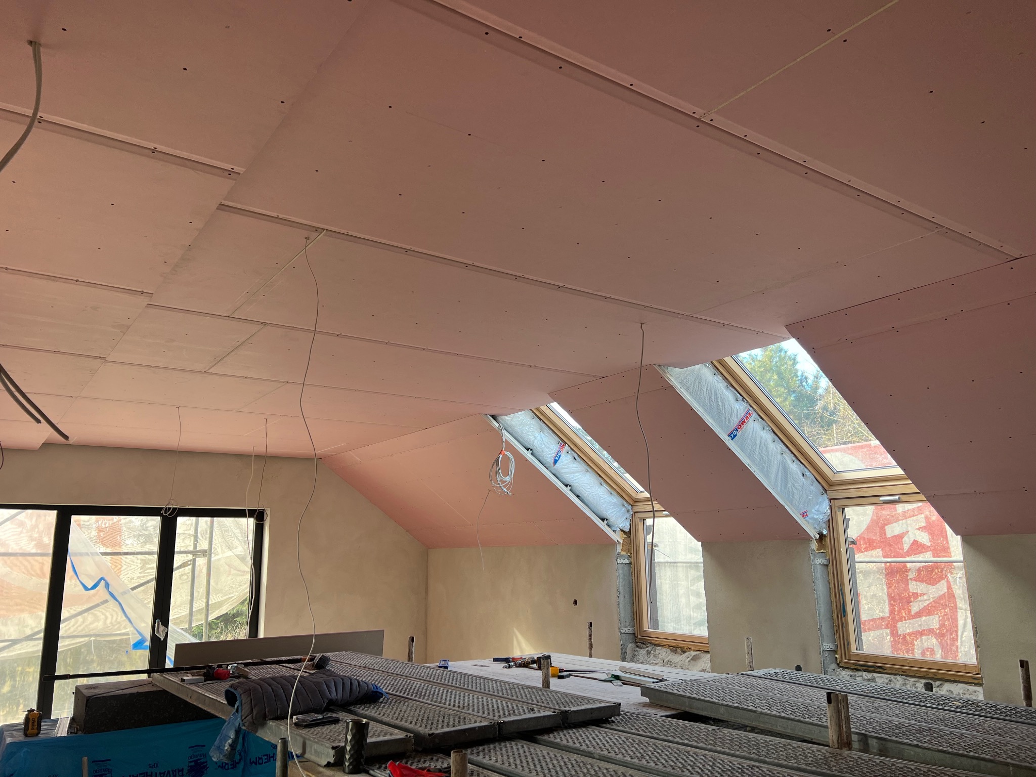 Poddasze w trakcie wykańczania: widoczne płyty kartonowo-gipsowe na skosach i suficie, okna dachowe z folią ochronną, metalowe rusztowania oraz narzędzia na podłodze.