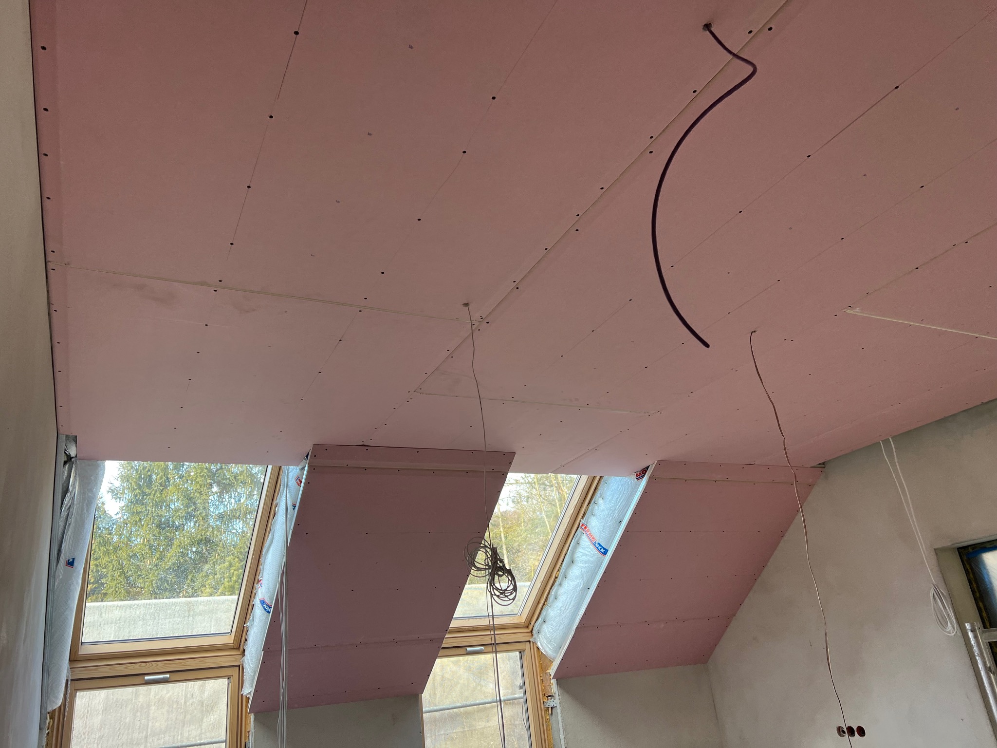 Poddasze z oknami dachowymi w trakcie montażu płyt gipsowo-kartonowych, widoczne przewody elektryczne i zabezpieczone okna folią.