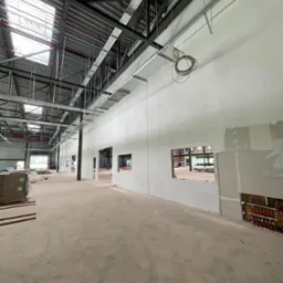 Wnętrze hali w trakcie prac wykończeniowych, widoczne ściany z częściowo położoną płytą karton-gips, odsłonięte instalacje grzewcze, metalowa konstrukcja dachu z oknami dachowymi.