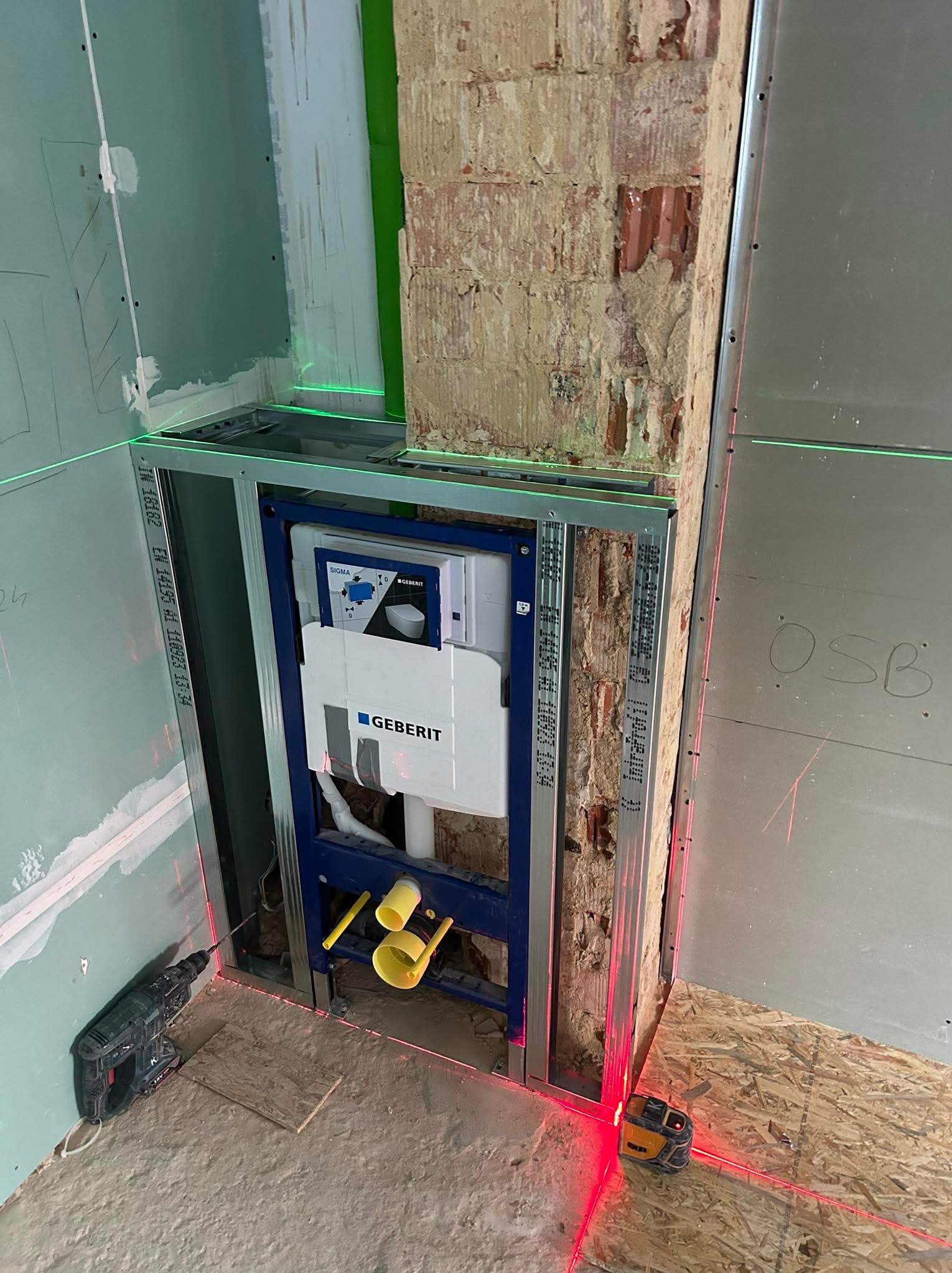 Stelaż podtynkowy Geberit zamontowany w konstrukcji z profili stalowych, widoczne płyty karton-gips i laser krzyżowy wyznaczający poziom, w tle fragment starej ceglanej ściany.