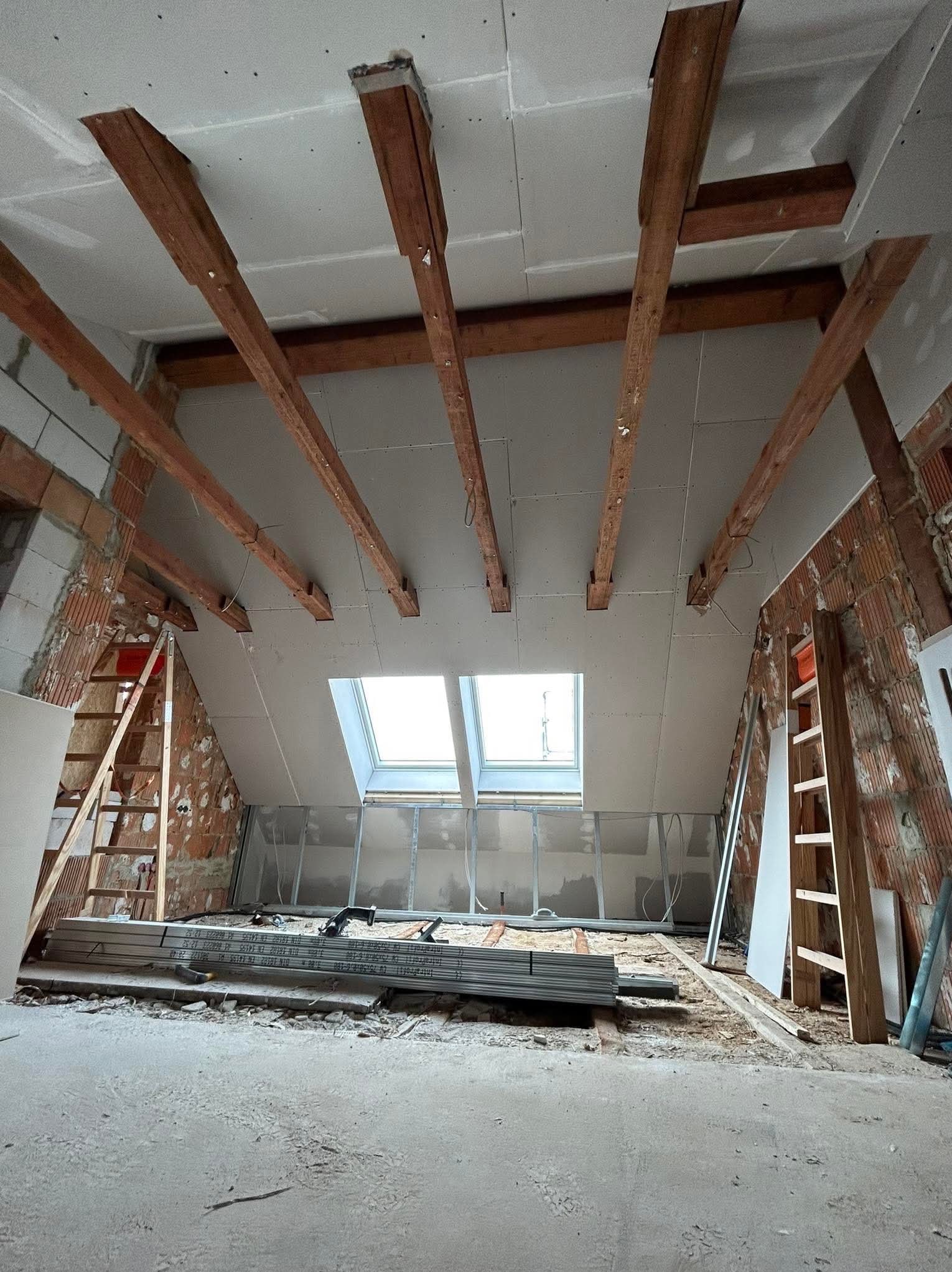 Montaż płyt kartonowo-gipsowych na poddaszu z widocznymi belkami stropowymi i oknem dachowym, w tle ściany z cegły i drabiny.