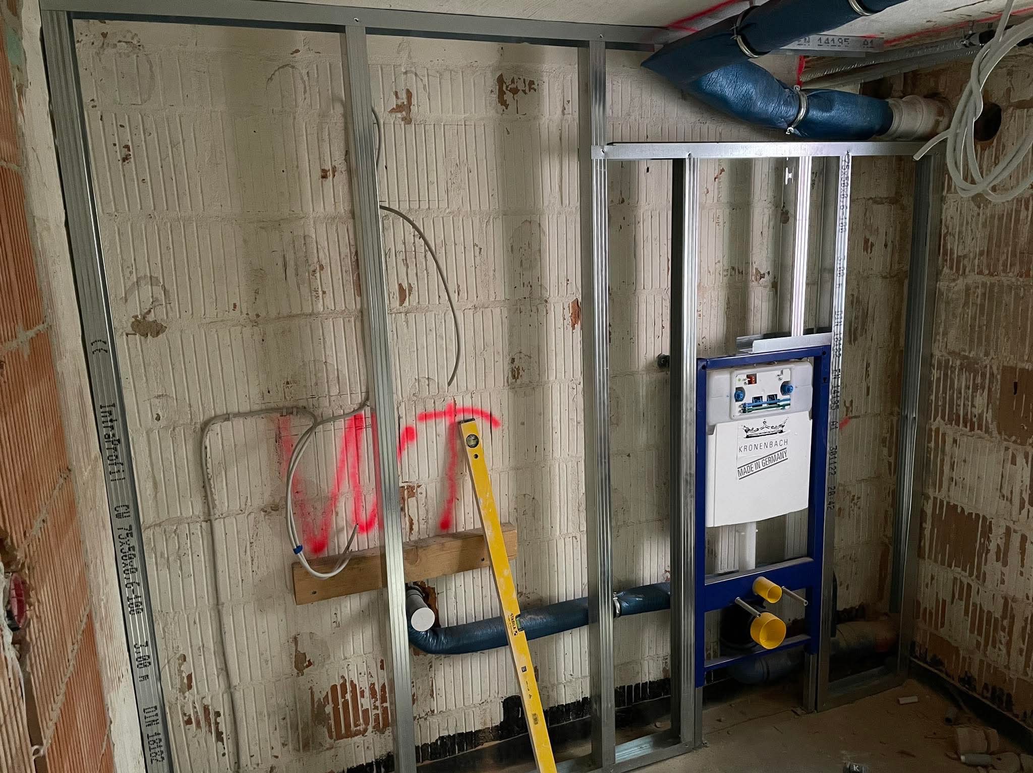 Montaż stelaża pod płytę karton gips w łazience z widocznymi instalacjami wodno-kanalizacyjnymi i poziomnicą Stabila opartą o ścianę z czerwonymi oznaczeniami.