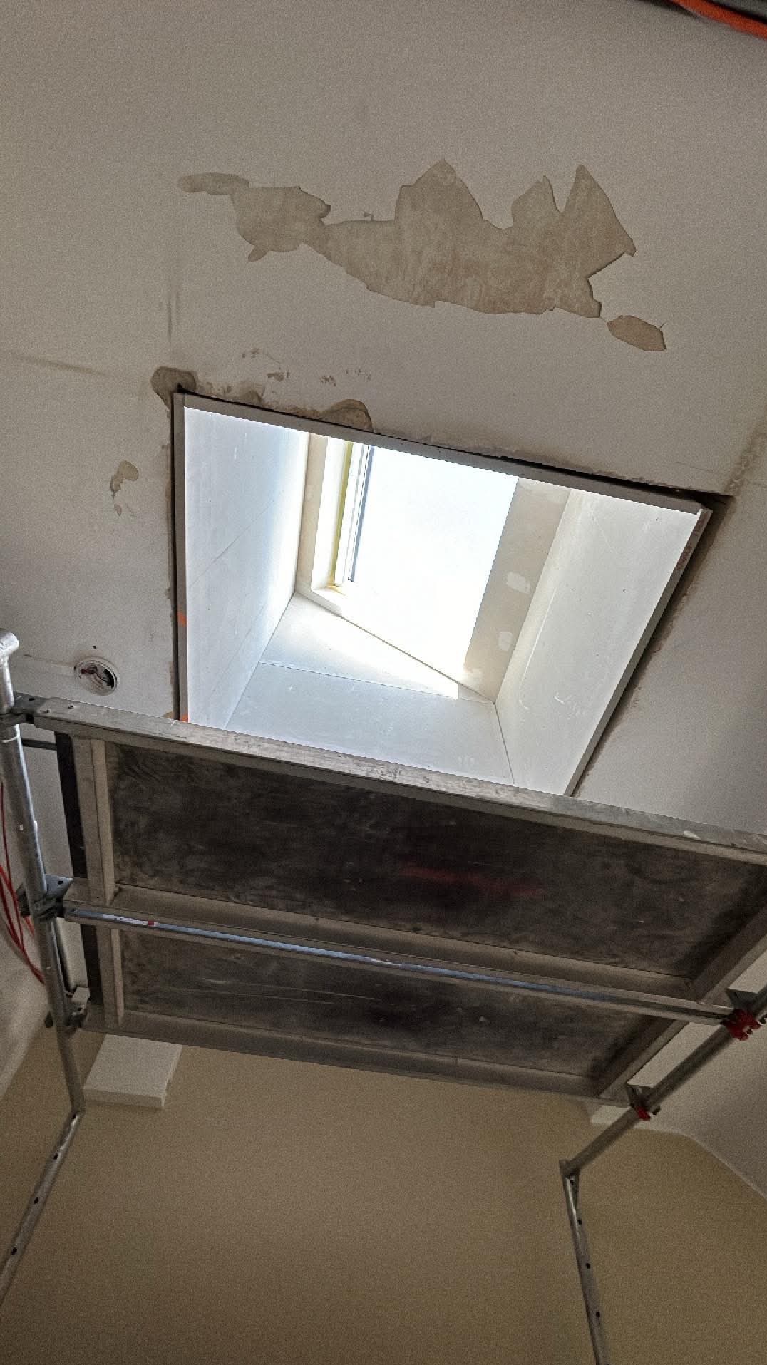 Montaż okna dachowego w trakcie prac wykończeniowych, widoczny fragment rusztowania, surowy tynk wokół otworu okiennego i częściowo przygotowane wnętrze wnęki okiennej z płyt kartonowo-gipsowych.