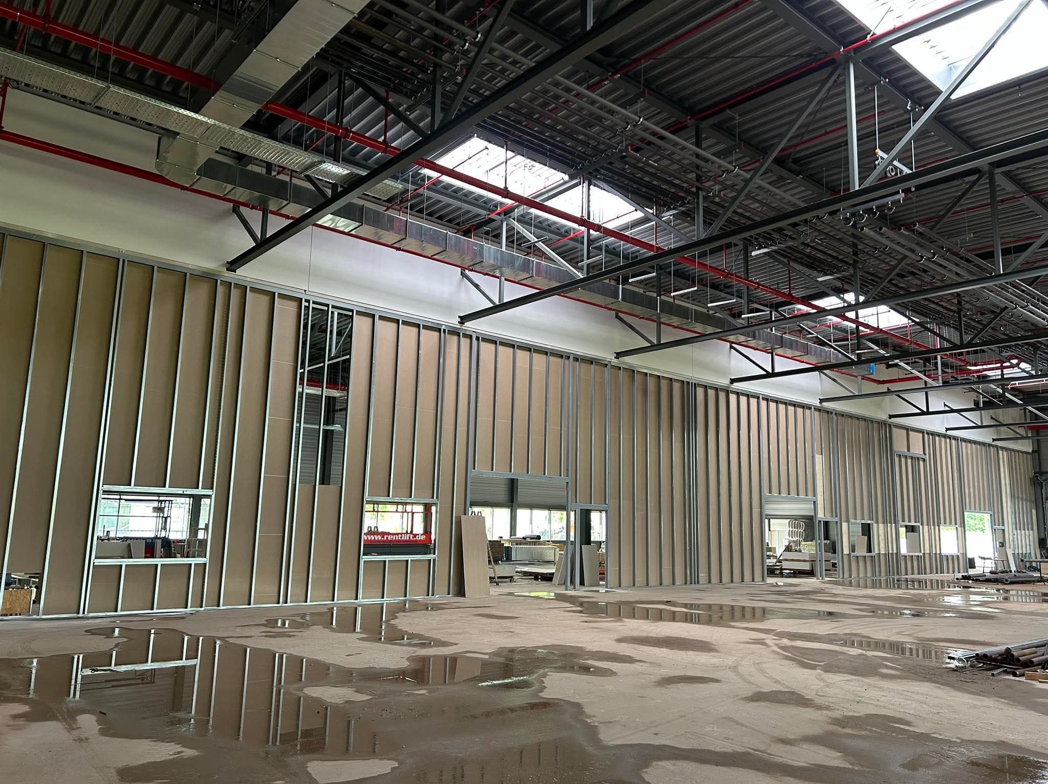 Wnętrze hali produkcyjnej w trakcie montażu ścian działowych z płyty kartonowo-gipsowej na metalowym stelażu, widoczne instalacje pod sufitem i mokra posadzka.