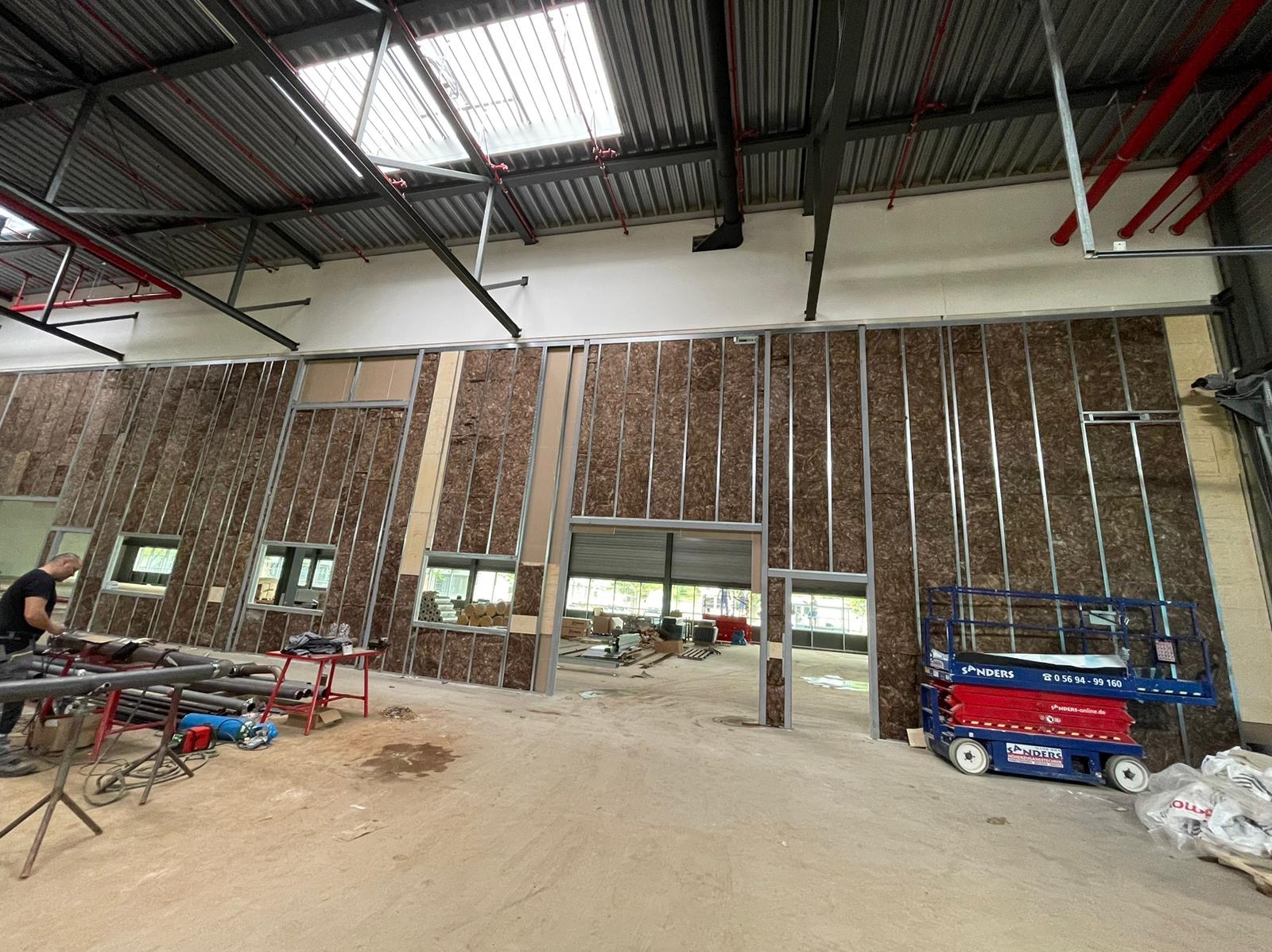 Montaż ścian działowych: metalowy szkielet z izolacją z wełny mineralnej, widoczne okna i brama garażowa w tle, podnośnik nożycowy i narzędzia w pomieszczeniu o surowym wykończeniu.