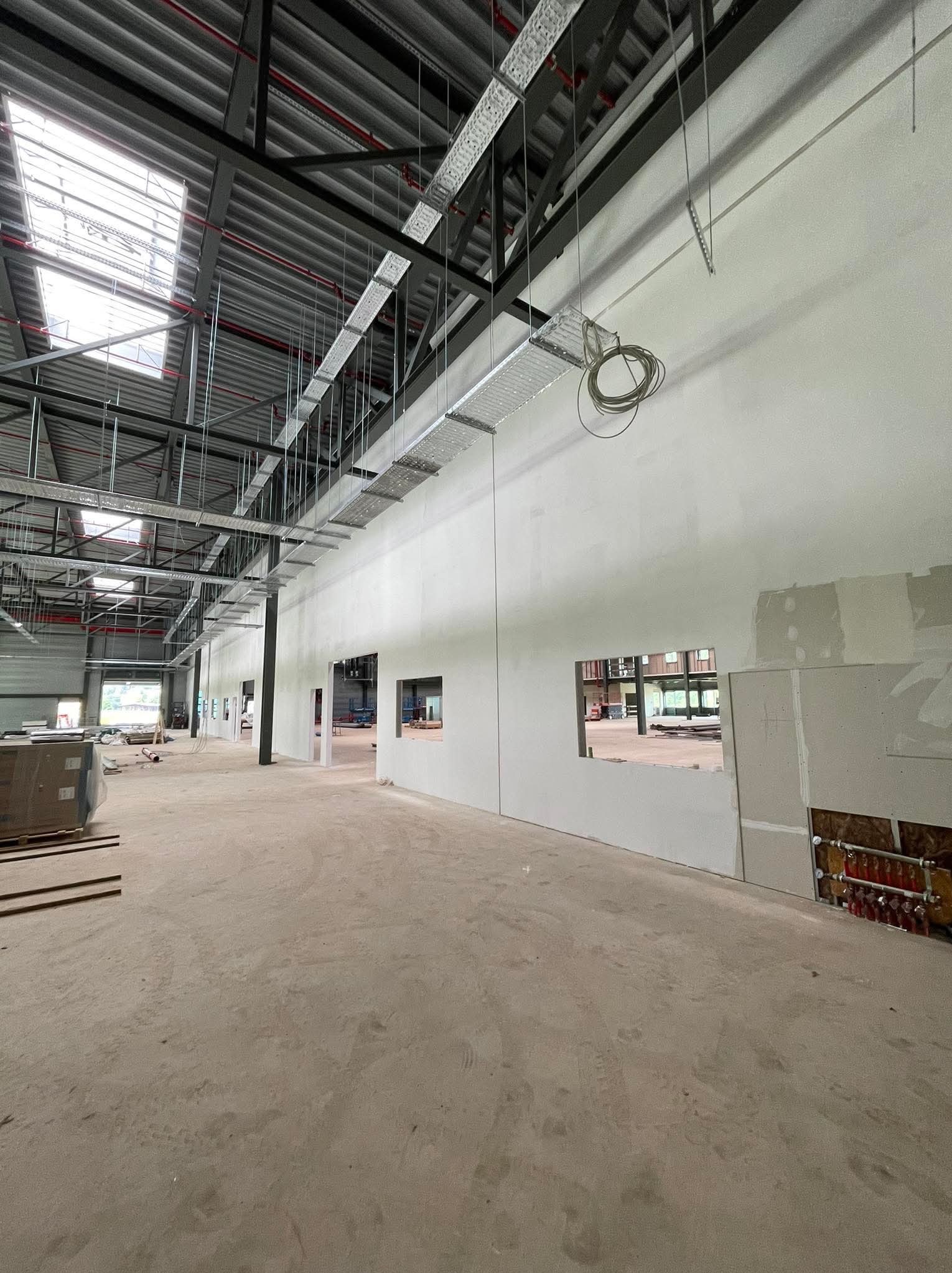 Wnętrze hali w trakcie prac wykończeniowych, widoczne ściany z częściowo położoną płytą karton-gips, odsłonięte instalacje grzewcze, metalowa konstrukcja dachu z oknami dachowymi.