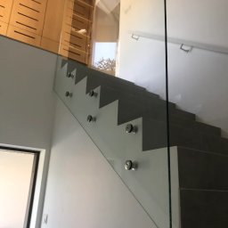 Alhen Artszkło Sp. z o.o. - Szklana balustrada zamocowana punktowo do schodów z szarymi stopniami, widok z dołu na górę, w tle drewniana szafa.