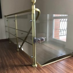 Alhen Artszkło Sp. z o.o. - Szklana barierka zabezpieczająca schody z metalowymi, złotymi słupkami i zamkiem na klucz, widok z dołu, na podłodze drewniane panele.