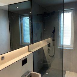 Alhen Artszkło Sp. z o.o. - Nowoczesna łazienka z kabiną prysznicową oddzieloną szkłem w czarnej ramie, podświetlanym blatem i podwieszanym bidetem w odcieniach szarości.