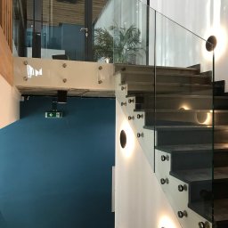 Alhen Artszkło Sp. z o.o. - Nowoczesne schody z szarobrunatnymi stopniami i szklaną balustradą mocowaną na metalowe śruby, oświetlone okrągłymi lampami ściennymi w minimalistycznym wnętrzu z błękitną ścianą i szklanymi drzwiami.