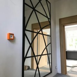 Alhen Artszkło Sp. z o.o. - Duże lustro w czarnej metalowej ramie z geometrycznym wzorem, zamontowane na białej ścianie w korytarzu, z widocznym pomarańczowym czujnikiem dymu po lewej stronie i otwartymi drzwiami w tle.