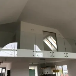 Szklana balustrada antresoli z białym panelem dolnym i metalowymi mocowaniami, widok z dołu na jasne wnętrze z oknem dachowym.