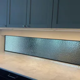 Panel szklany o fakturze lodu, zamontowany jako backsplash w kuchni, podświetlony listwą LED, widoczne górne szafki w kolorze grafitowym.