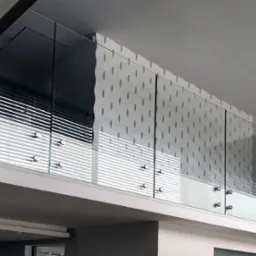 Szklana balustrada antresoli z widocznymi mocowaniami punktowymi, odbijająca światło z żaluzji i wzorzystej ściany.