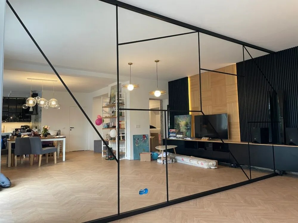 Nowoczesna szklana ścianka działowa z czarnymi profilami w salonie, widok na jadalnię i ścianę z lamelami.