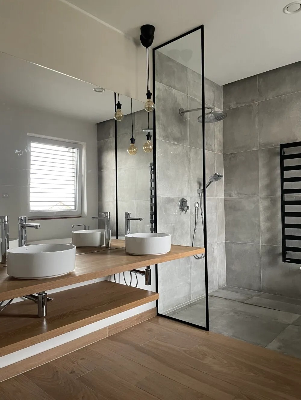 Nowoczesna łazienka z dwoma okrągłymi umywalkami na drewnianym blacie, szklaną kabiną prysznicową z czarną ramą i wiszącymi żarówkami Edison.