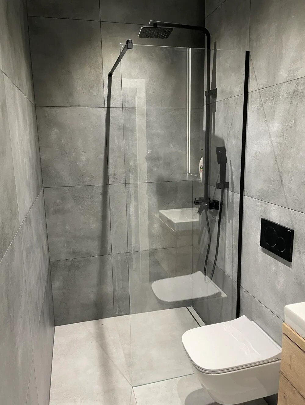 Nowoczesna łazienka z kabiną prysznicową typu walk-in oddzieloną szklaną ścianką z czarnymi okuciami, minimalistyczna toaleta podwieszana i umywalka z drewnianą szafką w tle, szare płytki...