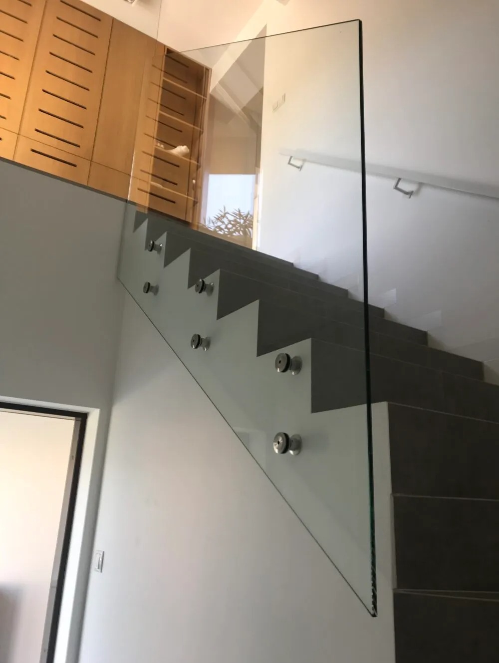 Szklana balustrada zamocowana punktowo do schodów z szarymi stopniami, widok z dołu na górę, w tle drewniana szafa.