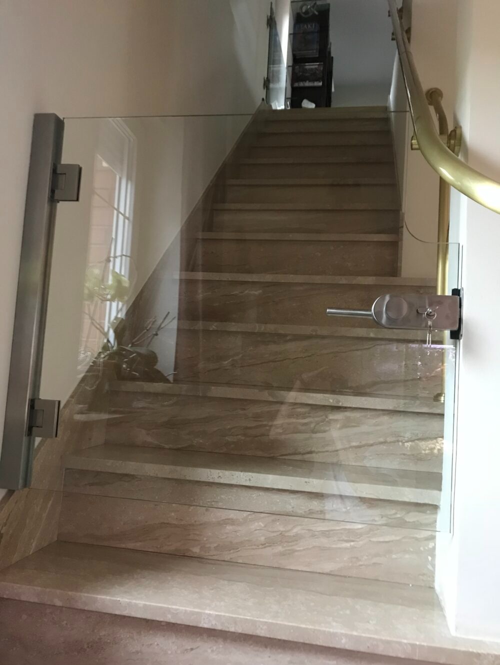 Szklana barierka zabezpieczająca schody z marmurowymi stopniami, zamontowana na zawiasach z metalowymi okuciami, z widoczną klamką i zamkiem na górze.