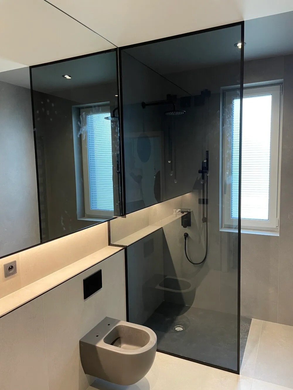 Nowoczesna łazienka z kabiną prysznicową oddzieloną szkłem w czarnej ramie, podświetlanym blatem i podwieszanym bidetem w odcieniach szarości.