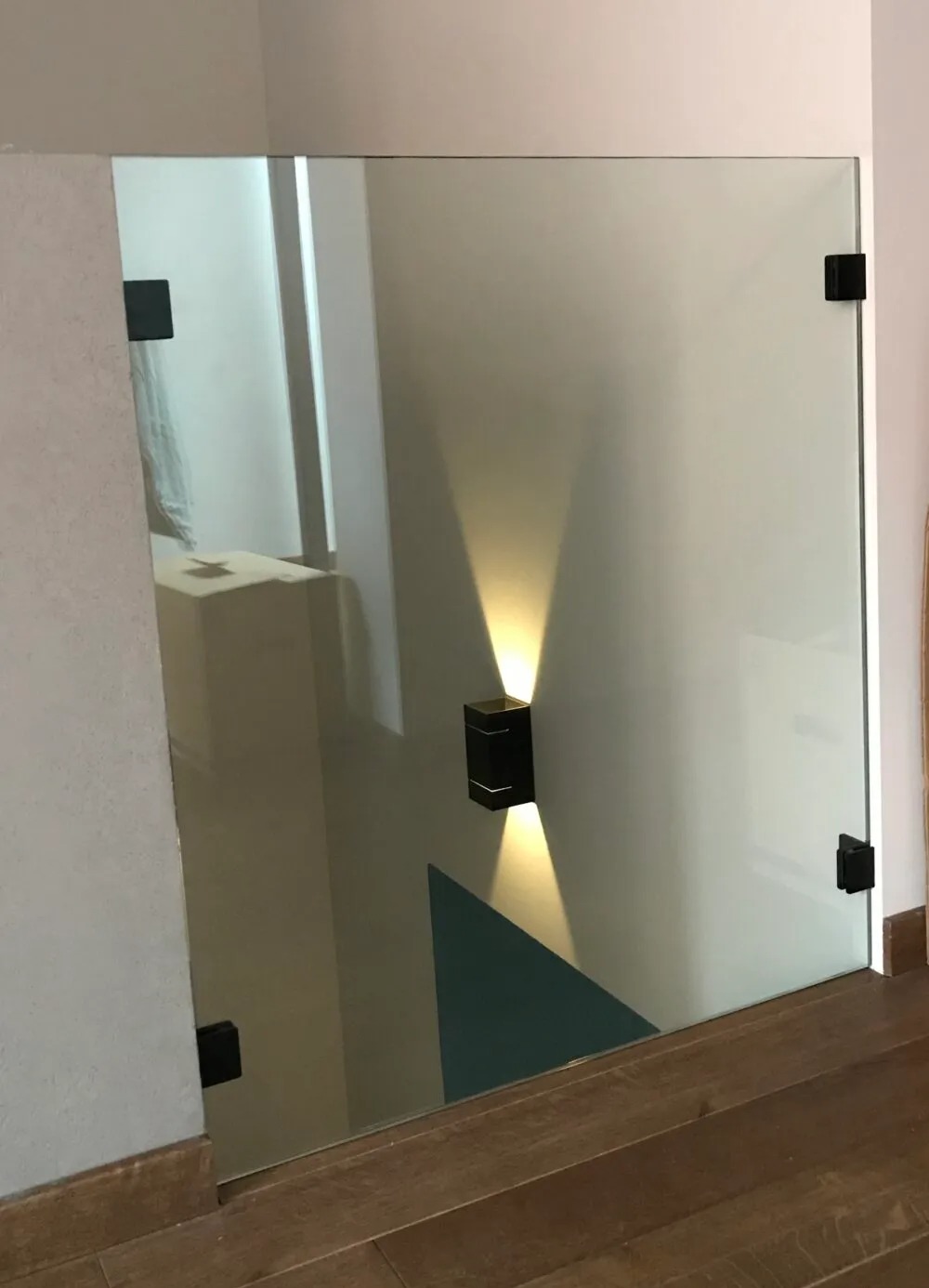 Szklana ściana działowa z matowego szkła z czarnymi okuciami, oświetlona kinkietem z dwoma strumieniami światła, widoczny fragment podłogi z drewnianych paneli.