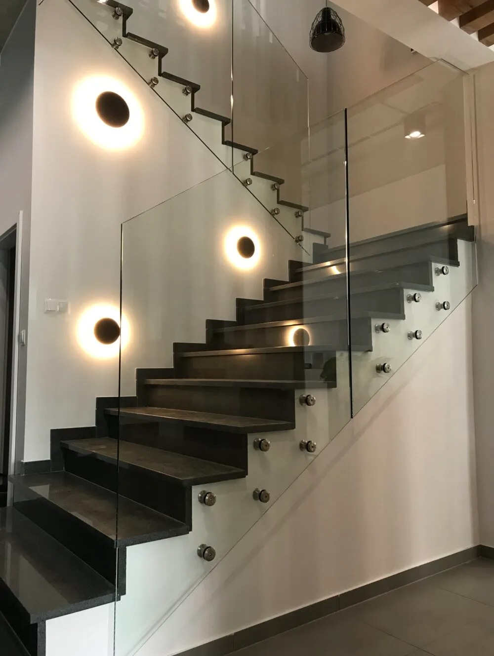 Nowoczesne schody z ciemnego kamienia z balustradą ze szkła mocowaną na metalowych elementach, podświetlone okrągłymi lampami ściennymi.