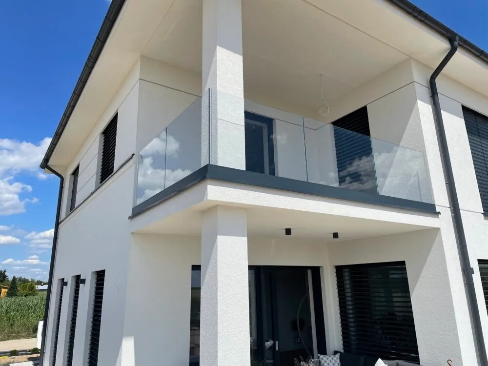 Nowoczesny balkon z balustradą ze szkła bezramowego na tle białej fasady domu i błękitnego nieba.