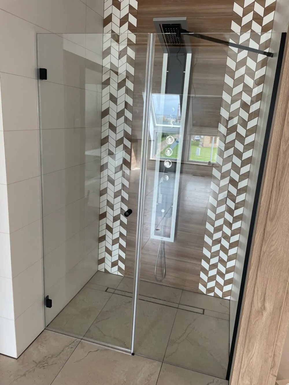 Kabina prysznicowa ze szklanymi drzwiami, wykończona kafelkami w chevronowy wzór, z widocznym odpływem liniowym i deszczownicą.