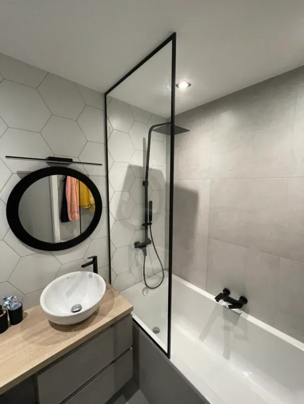 Nowoczesna łazienka z wanną i szklanym parawanem, białą umywalką nablatową na szarej szafce z drewnianym blatem, czarną armaturą i lustrem w okrągłej czarnej ramie na ścianie z heksagonalnymi...