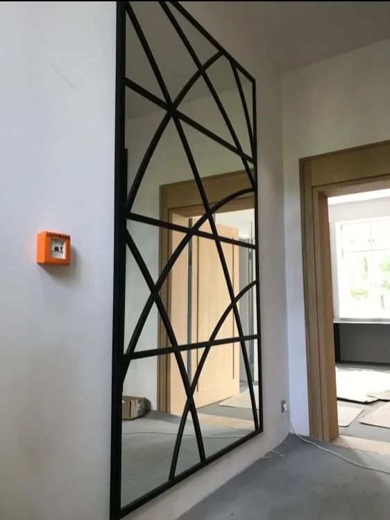 Duże lustro w czarnej metalowej ramie z geometrycznym wzorem, zamontowane na białej ścianie w korytarzu, z widocznym pomarańczowym czujnikiem dymu po lewej stronie i otwartymi drzwiami w tle.