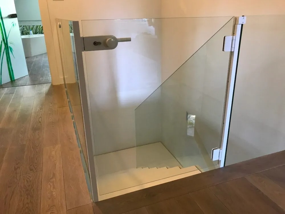 Szklane drzwi zabezpieczające schody wewnątrz domu, z widocznym zamkiem z boku i białymi zawiasami, zamontowane na białej ramie.