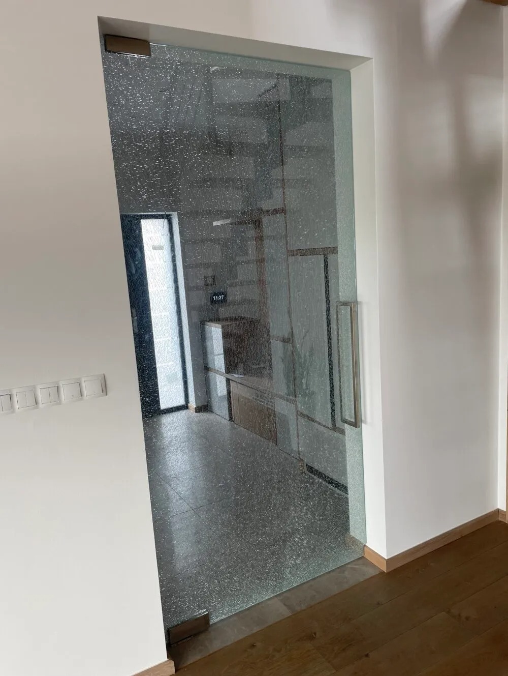 Przesuwne drzwi ze szkła ornamentowego z metalowym uchwytem, widok na przedpokój z szarą podłogą i oknem.