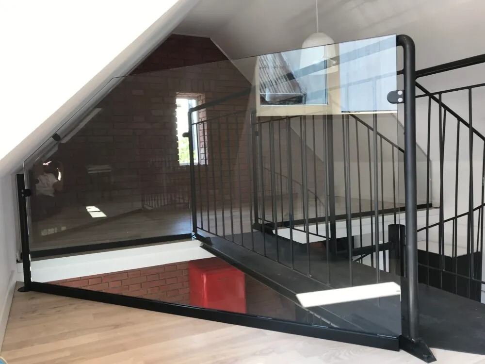 Szklana balustrada z czarnymi elementami mocującymi, zabezpieczająca schody spiralne na poddaszu z ceglaną ścianą i oknem.