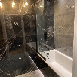 Jarosław Nitychoruk - Nowoczesna łazienka z czarnymi płytkami imitującymi marmur, białą wanną i czarną armaturą, oświetlona wiszącymi żarówkami bez kloszy. Widoczne odbicie osoby robiącej zdjęcie w szklanej ściance...