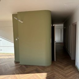Jarosław Nitychoruk - Wnętrze po remoncie z parkietem ułożonym w jodełkę, ścianą w odcieniu oliwkowej zieleni i białymi listwami przypodłogowymi. Widoczne niedokończone podłączenie przewodów elektrycznych.