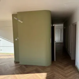 Wnętrze po remoncie z parkietem ułożonym w jodełkę, ścianą w odcieniu oliwkowej zieleni i białymi listwami przypodłogowymi. Widoczne niedokończone podłączenie przewodów elektrycznych.